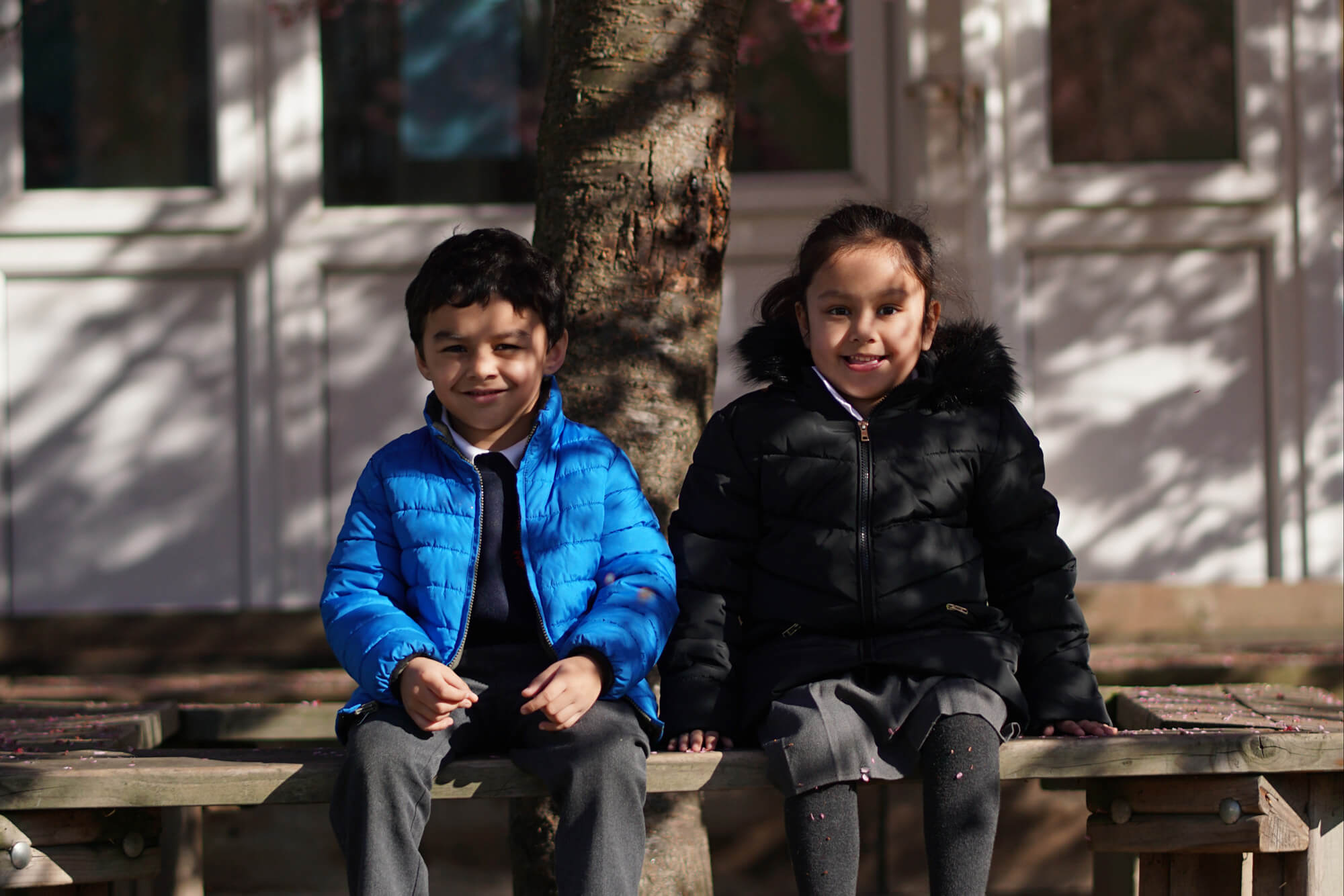 Infants – Two students sitting on bench under tree-crop
